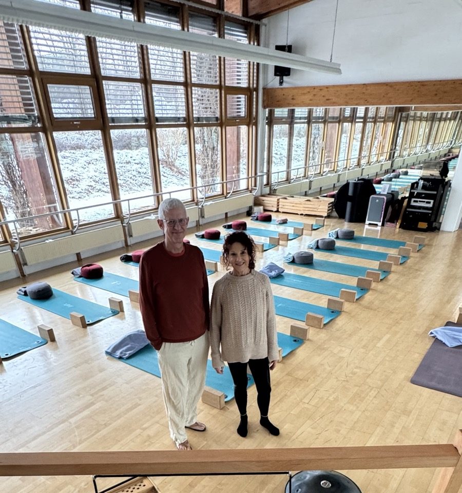 Ausgelegte Yogamatten im Kursraum vor den Panoramafenstern, zwei Menschen im Gespräch